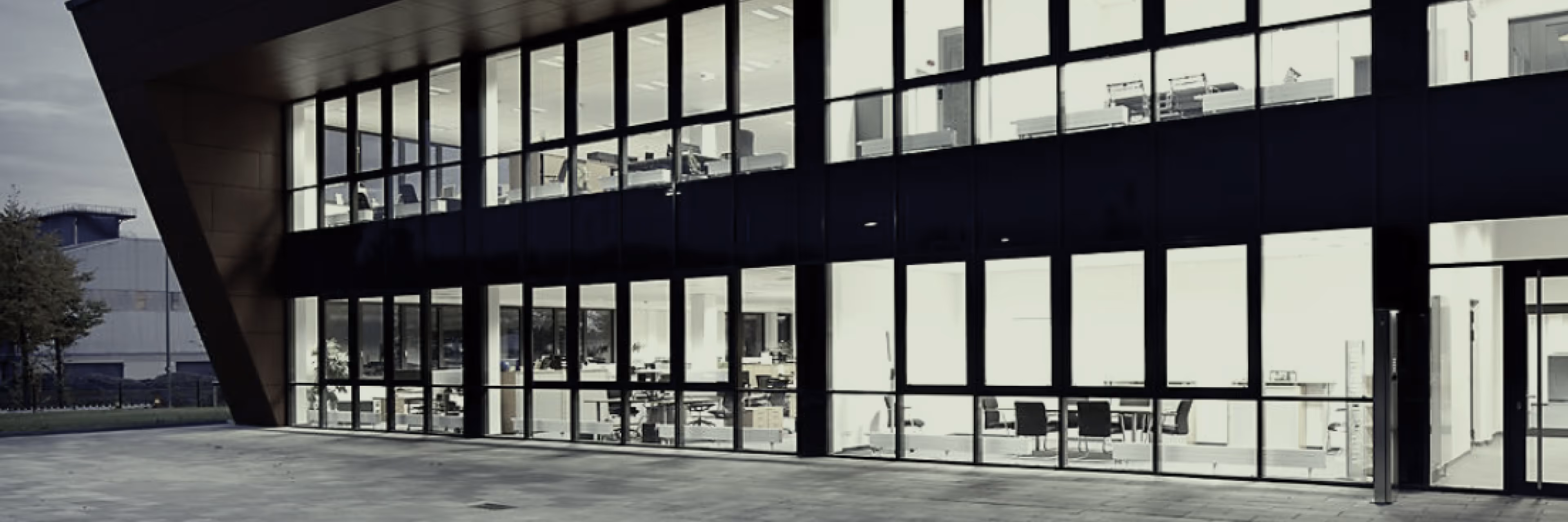 Modernes Bürogebäude mit großen Glasfenstern und Blick auf Innenräume mit Schreibtischen und Stühlen.
