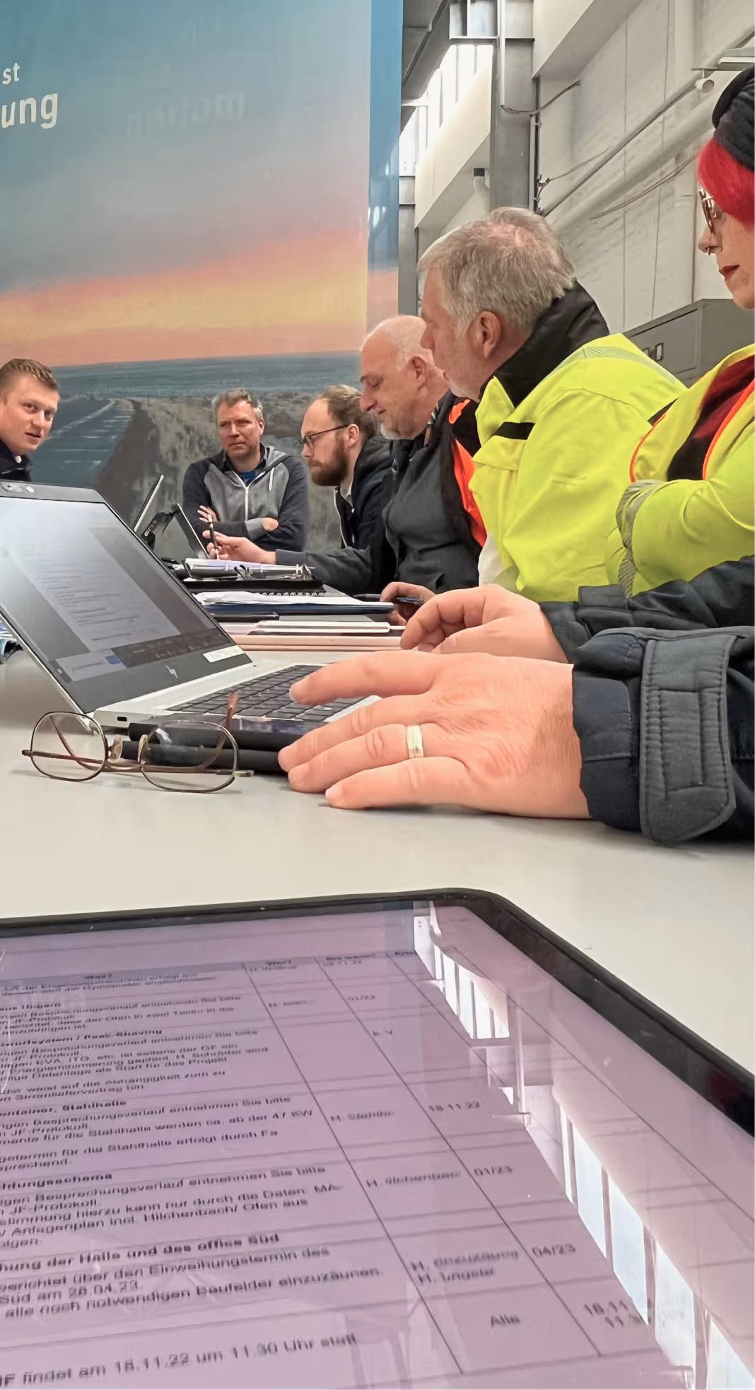 Sechs Personen in Arbeitskleidung sitzen bei einem Meeting mit Laptops und Dokumenten an einem Tisch.