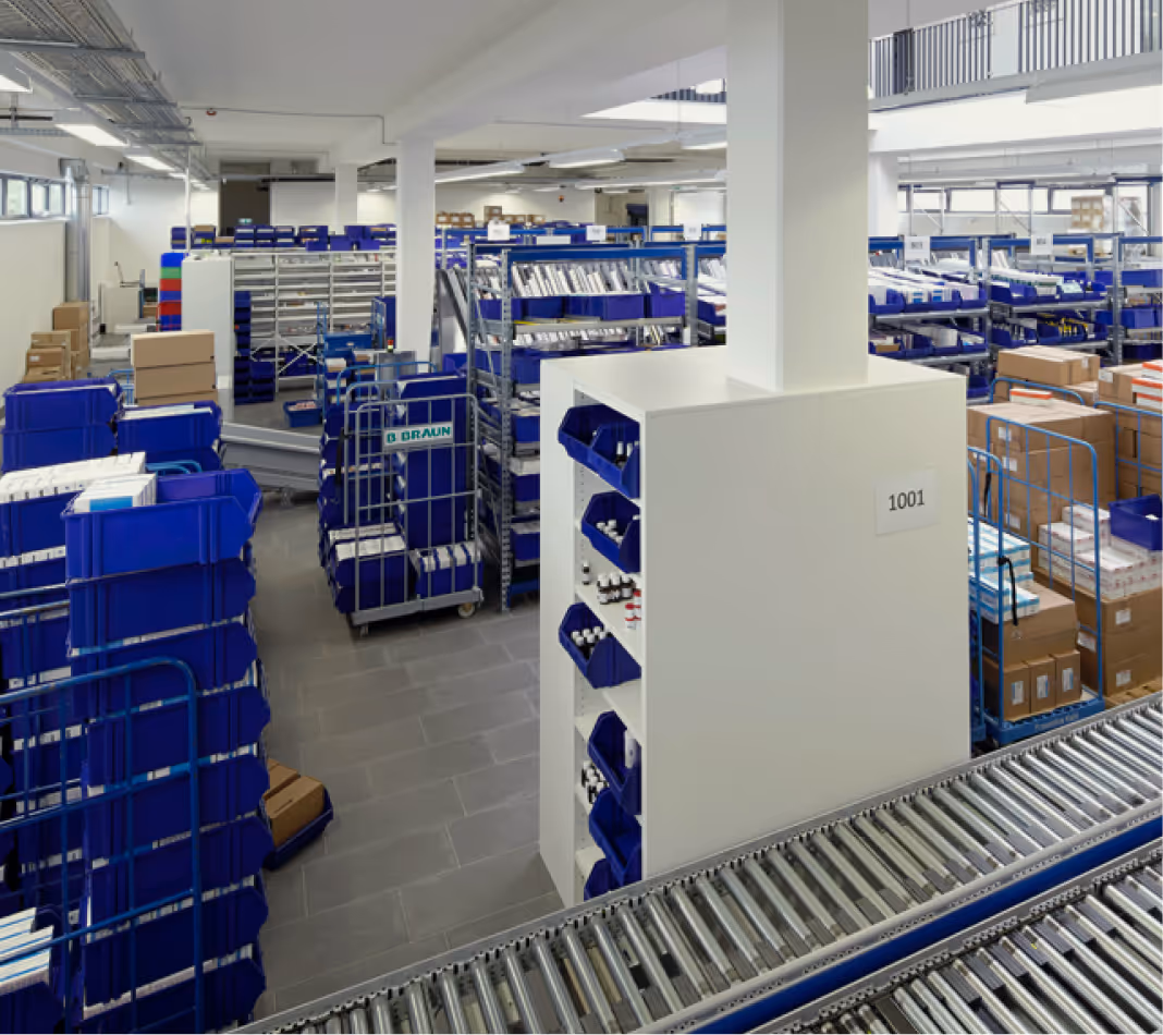 Großes Lager mit blauen Behältern, Kartons und Metallregalen in einer hellen Halle mit Rollbahn.