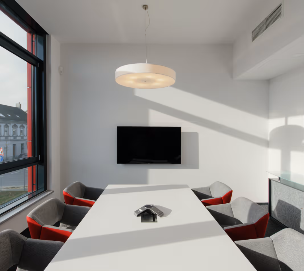 Moderner Konferenzraum mit weißem Tisch, grauen Stühlen mit roten Akzenten und Wand-TV.