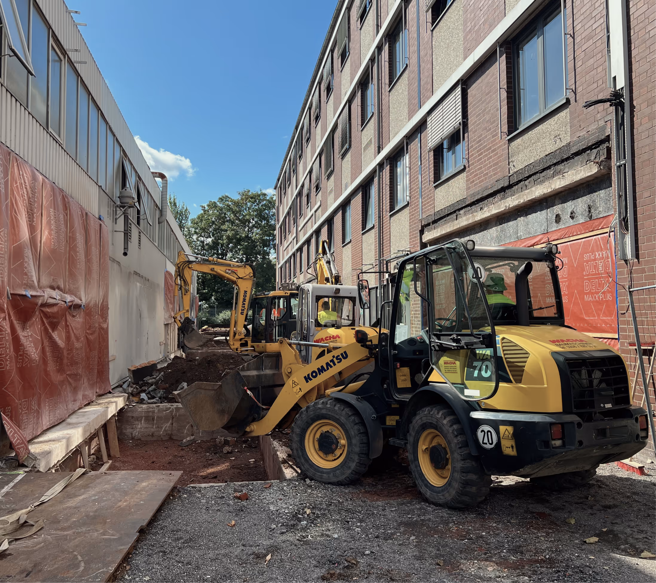 Baustelle zwischen Industriegebäuden mit Radlader und Bagger bei Erdarbeiten.