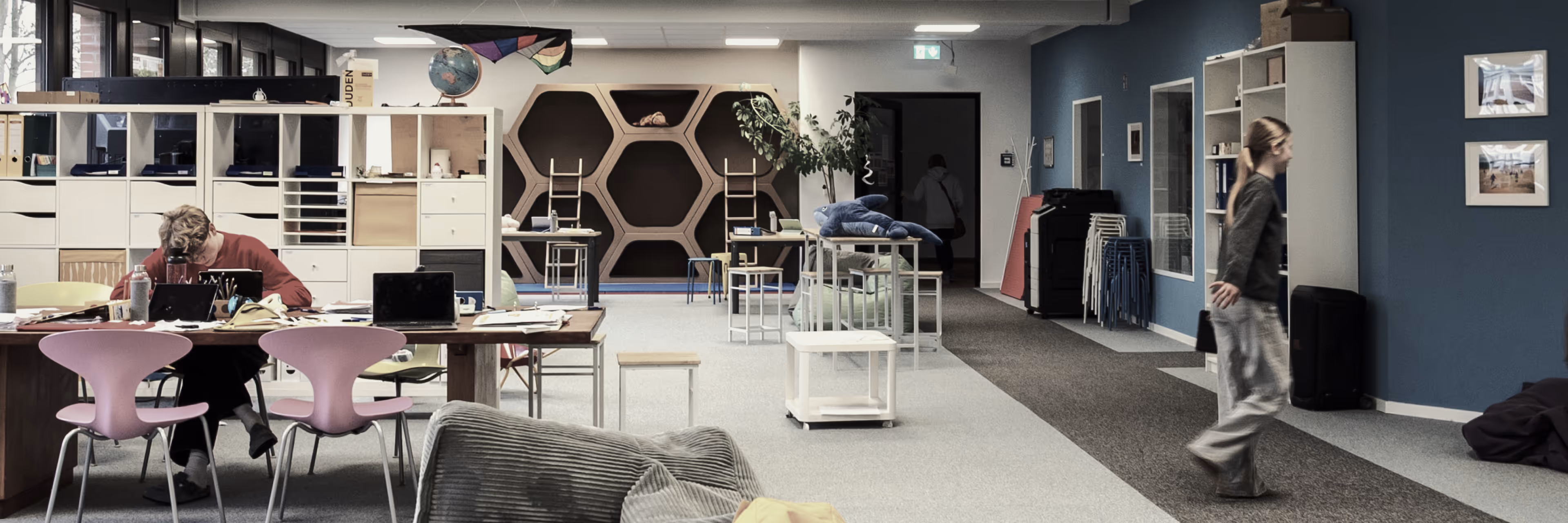 Moderner Gemeinschaftsarbeitsraum mit Tischen, Stühlen, Regalen und Personen beim Arbeiten und Bewegen.