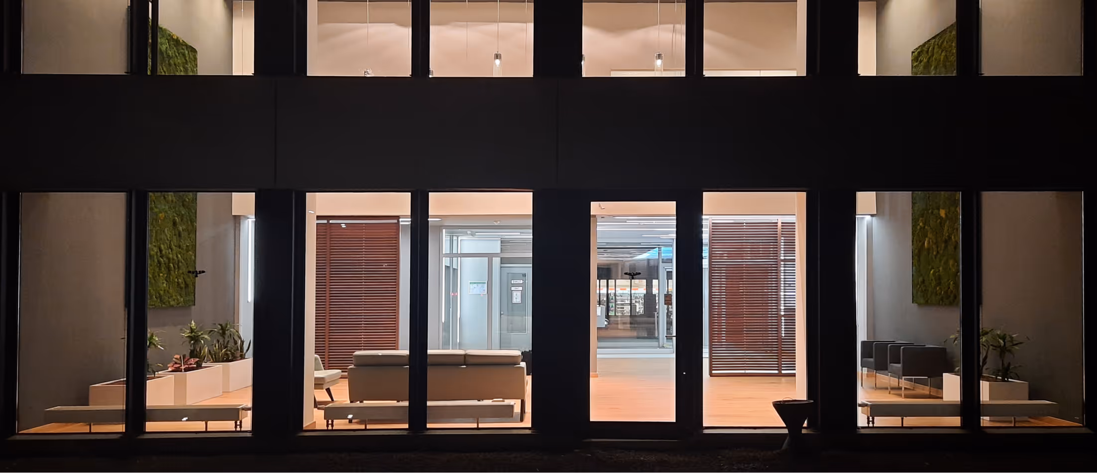 Blick durch Fenster auf moderne Lobby mit Sofas, Pflanzen und Holzboden bei Nacht.