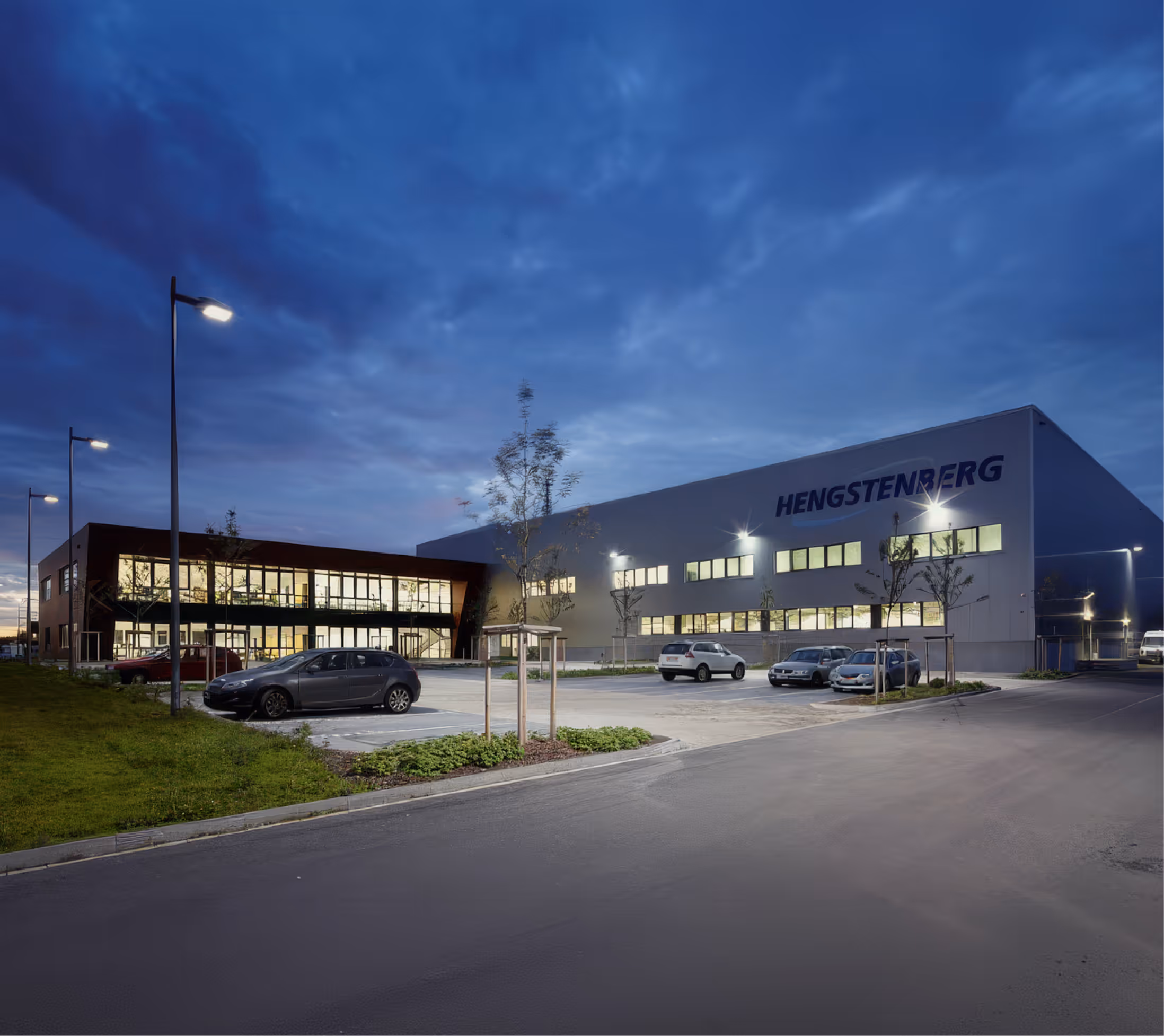 Beleuchtetes Büro- und Industriegebäude mit Parkplatz und Autos bei Abenddämmerung.