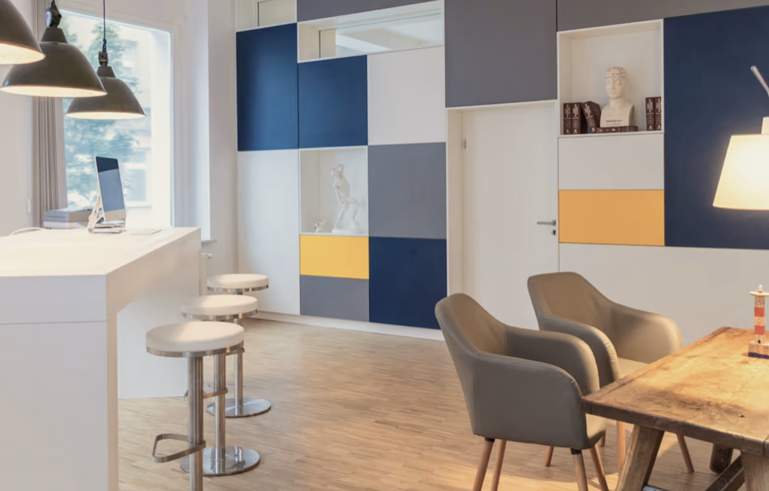 Modern Büro mit weißen Barhockern, grauen Sesseln, Holztisch und Wandregalen in Blau und Gelb.