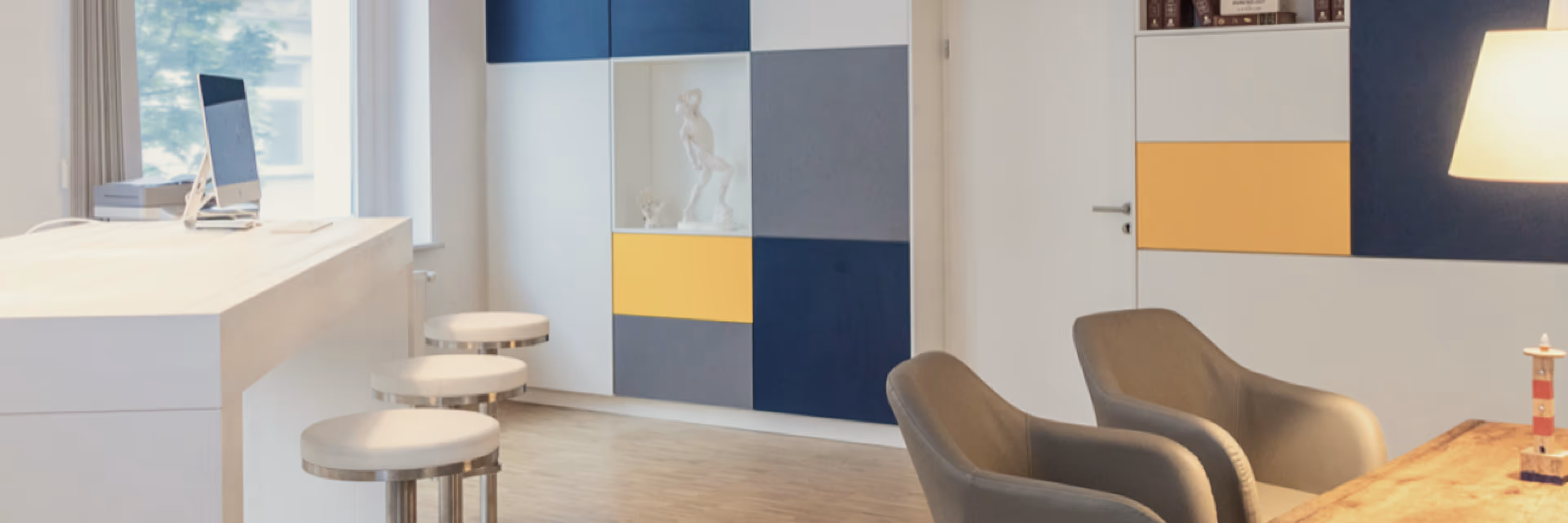 Modernes Büro mit weißen Hockern, grauen Sesseln, einem Schreibtisch, bunten Schränken und Lampenlicht.