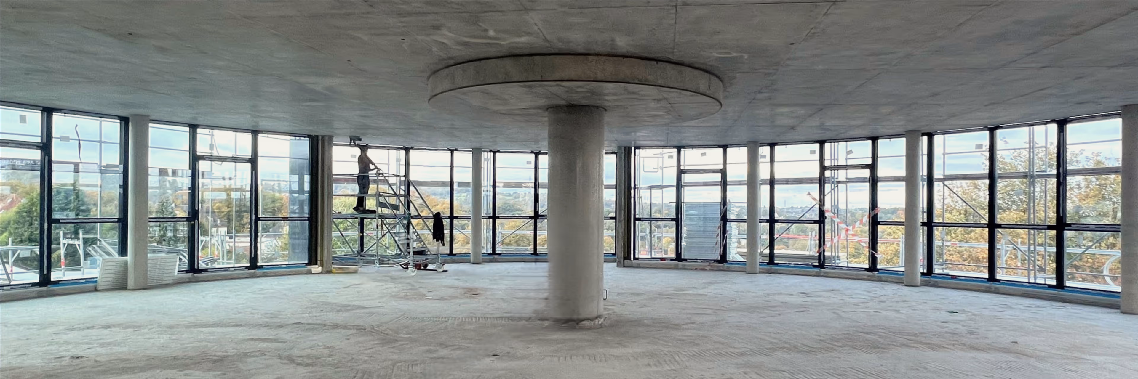 Offener Innenraum mit großem runden Betonsäulenelement und Fensterfront, Bauarbeiter auf Gerüst.
