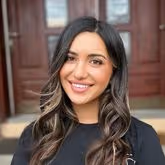 Smiling woman with long wavy dark hair and light highlights, wearing a black top, standing in front of a wooden door.