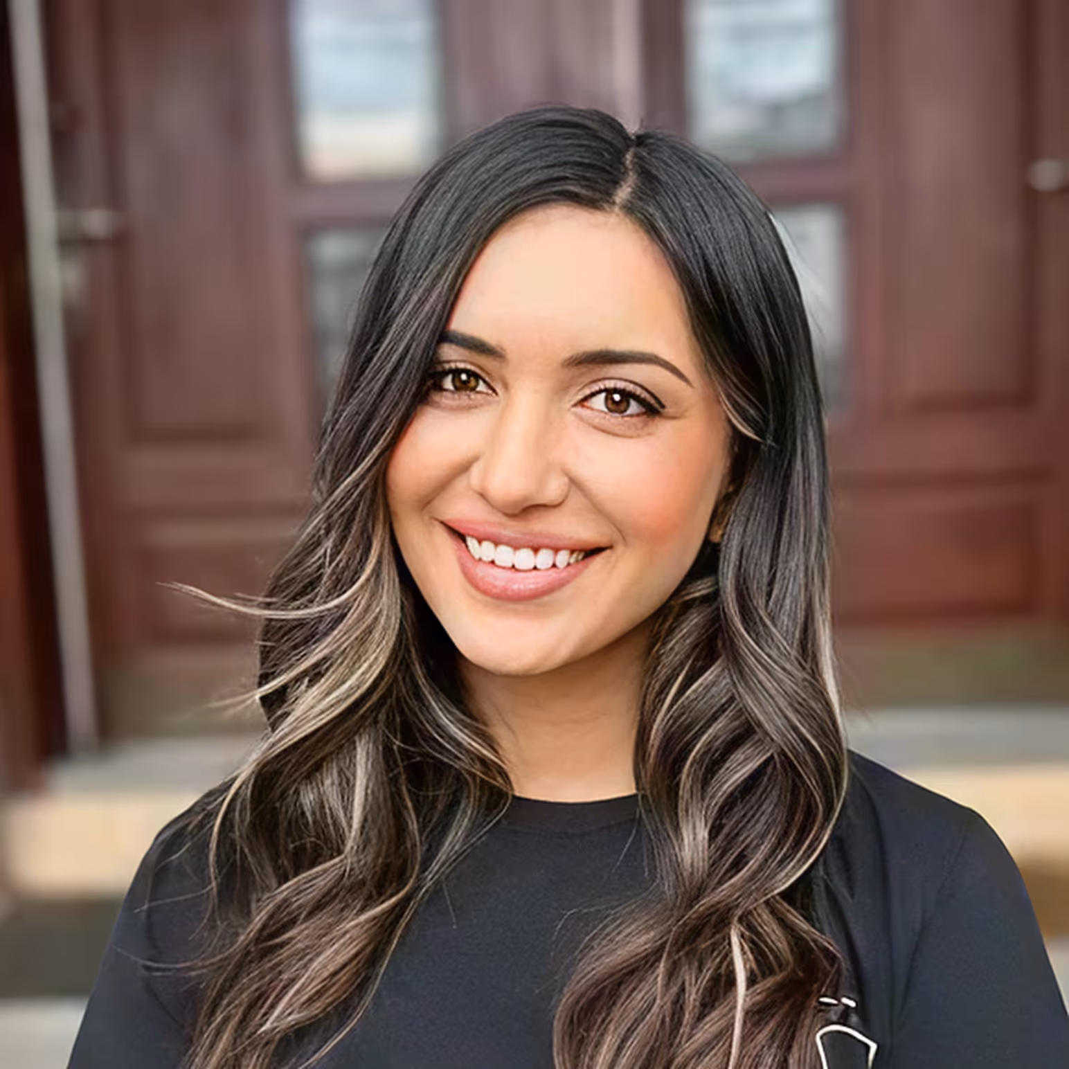 Smiling woman with long, wavy black hair with light highlights wearing a black top in front of a wooden door background.