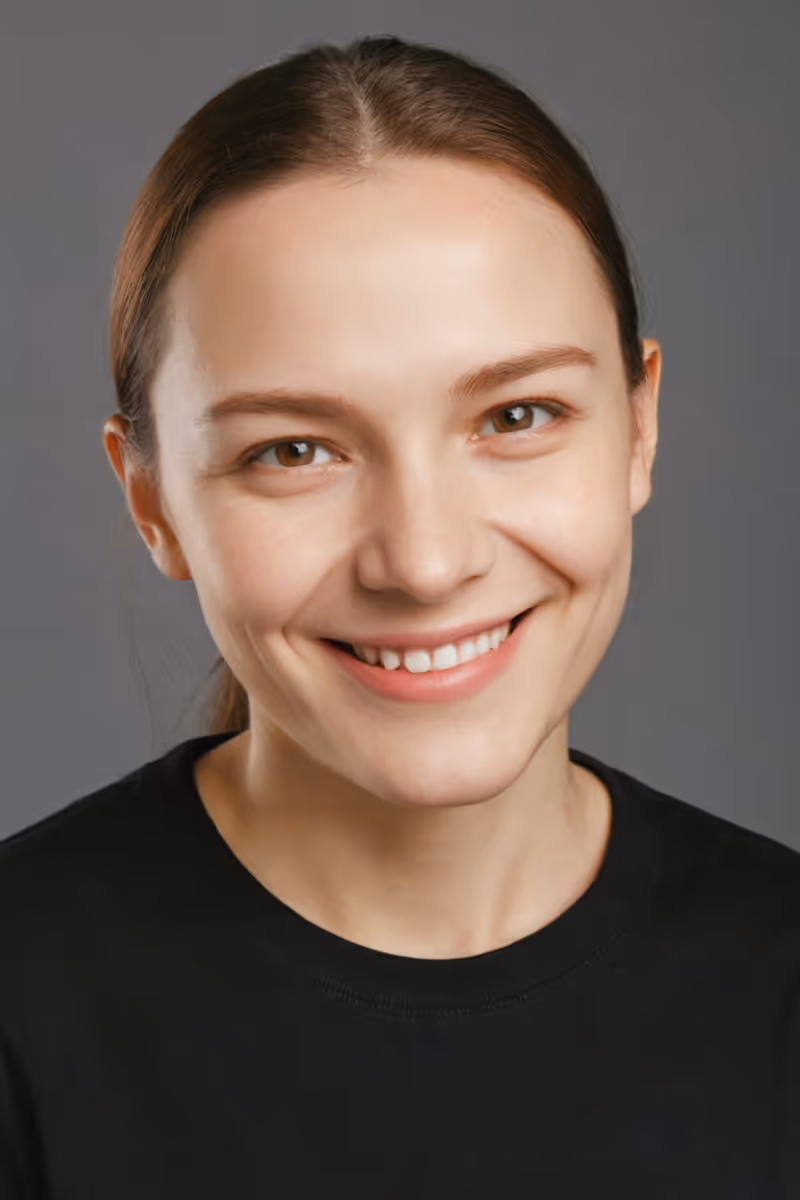 Smiling young woman with brown hair pulled back wearing a black shirt against a gray background.