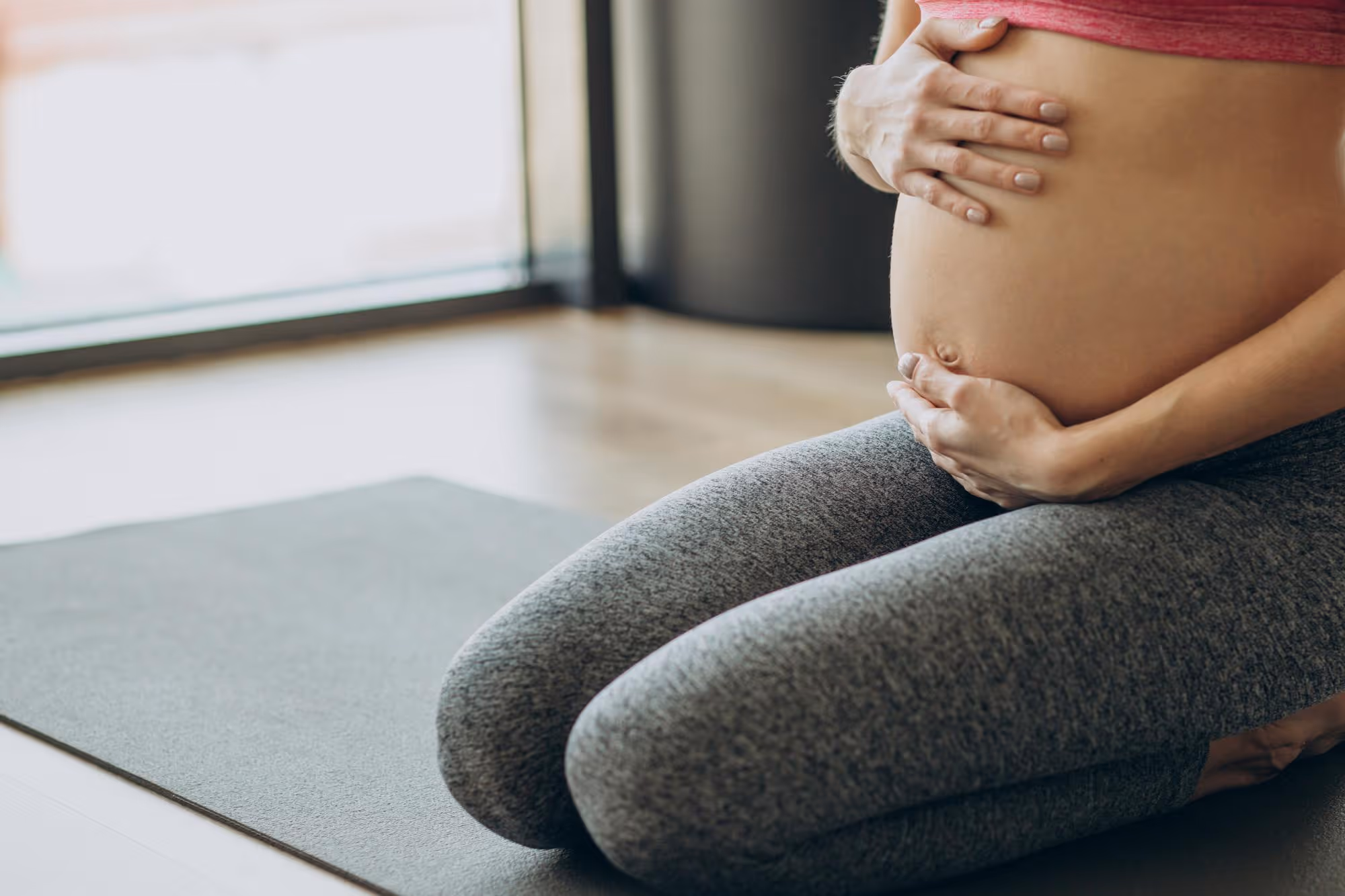 Close-up of pregnant woman kneeling on a yoga mat, holding her belly with both hands.