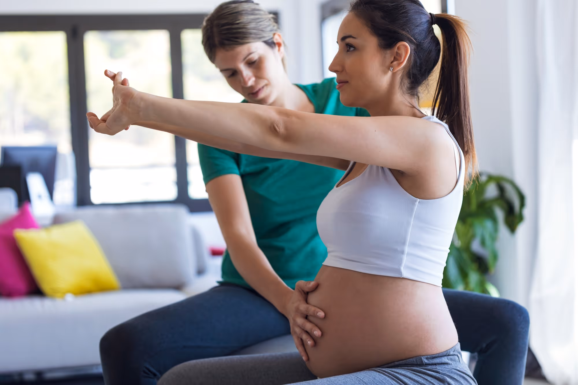 Pregnant woman in workout clothes stretching her arm forward while a female instructor supports her abdomen during exercise indoors.
