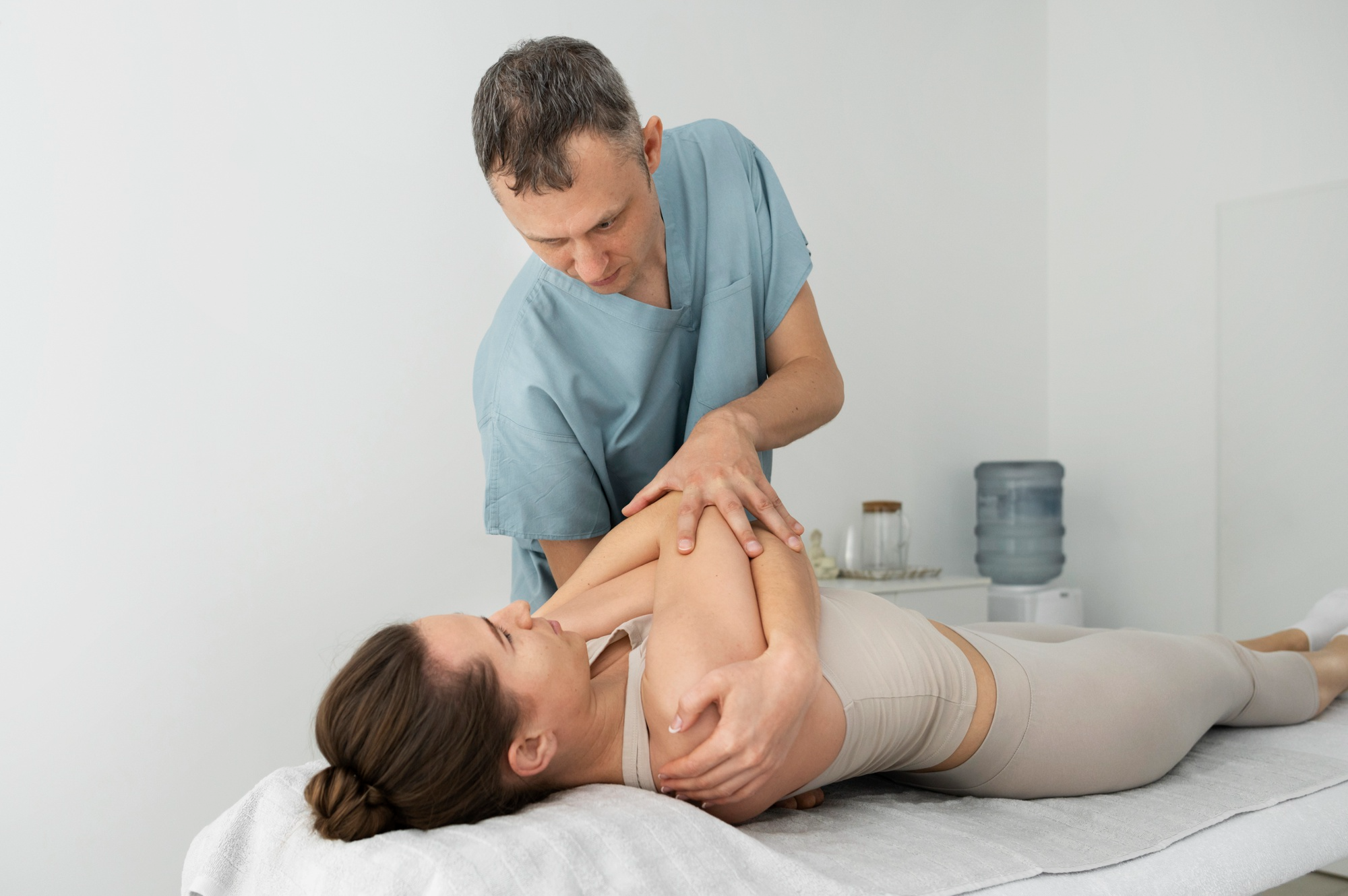 Physical therapist in blue scrubs stretching the arm of a woman lying on a treatment table in a clinical setting.