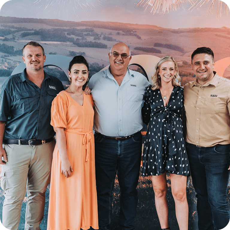 Five people standing together smiling in front of a scenic backdrop, three men wearing shirts with 'saai' logos and two women in dresses.
