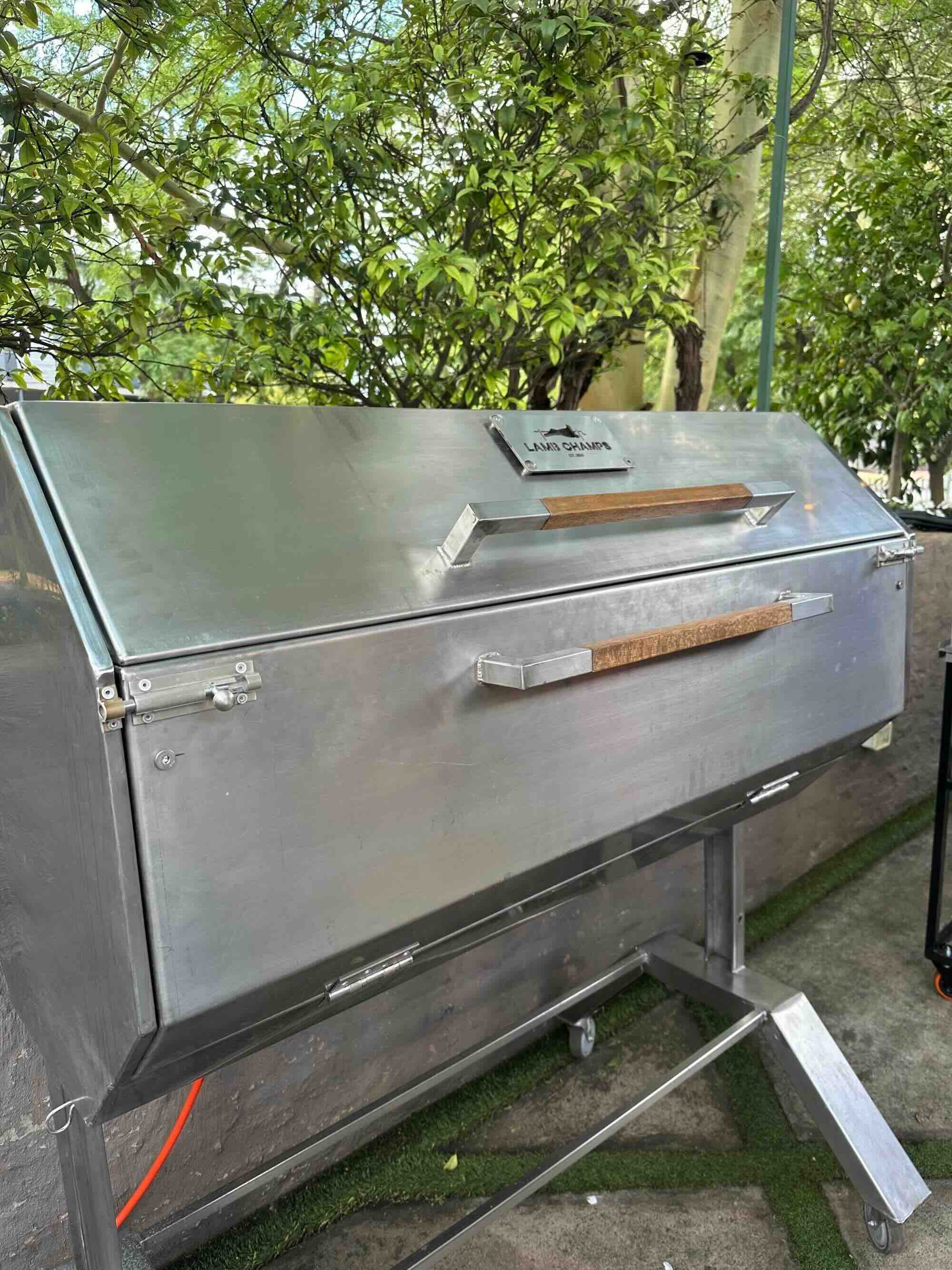 Large stainless steel grill with wooden handles labeled Lamb Champs outdoors near plants.