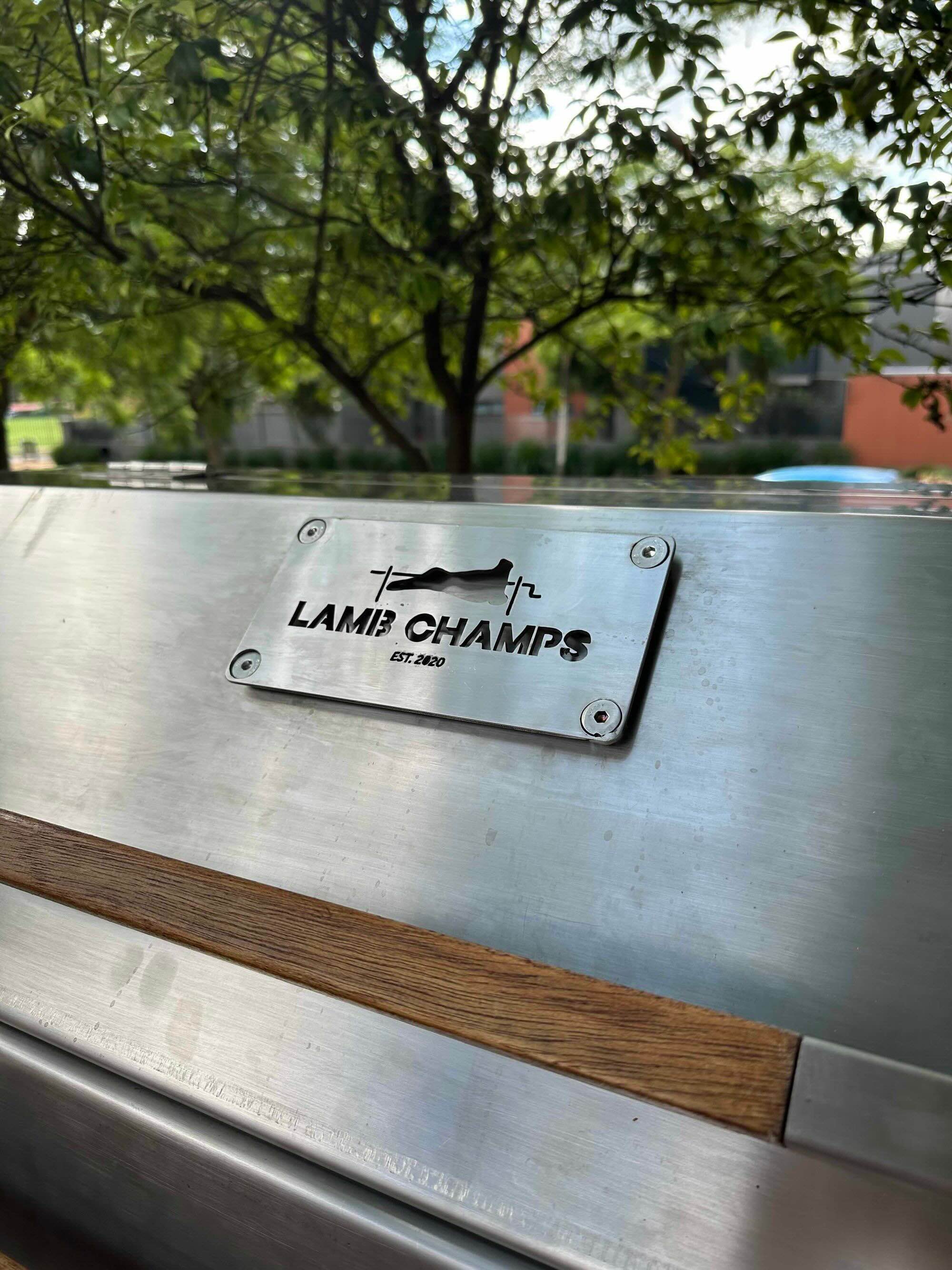 Metal plaque on grill lid reading 'Lamb Champs Est. 2020' with a lamb and spit graphic.