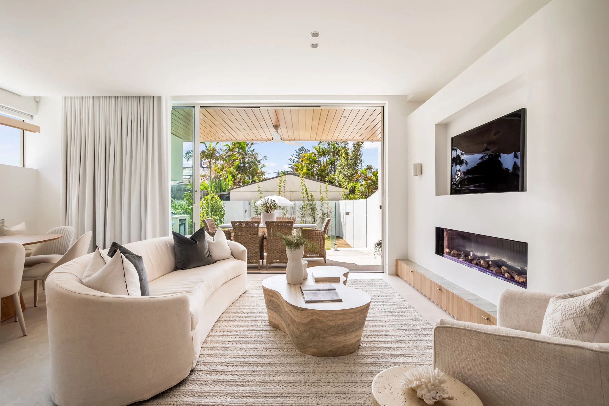 Bright modern living room with curved beige sofa, stone coffee table, armchair, wall-mounted TV and fireplace, overlooking an outdoor dining area through large sliding glass doors.