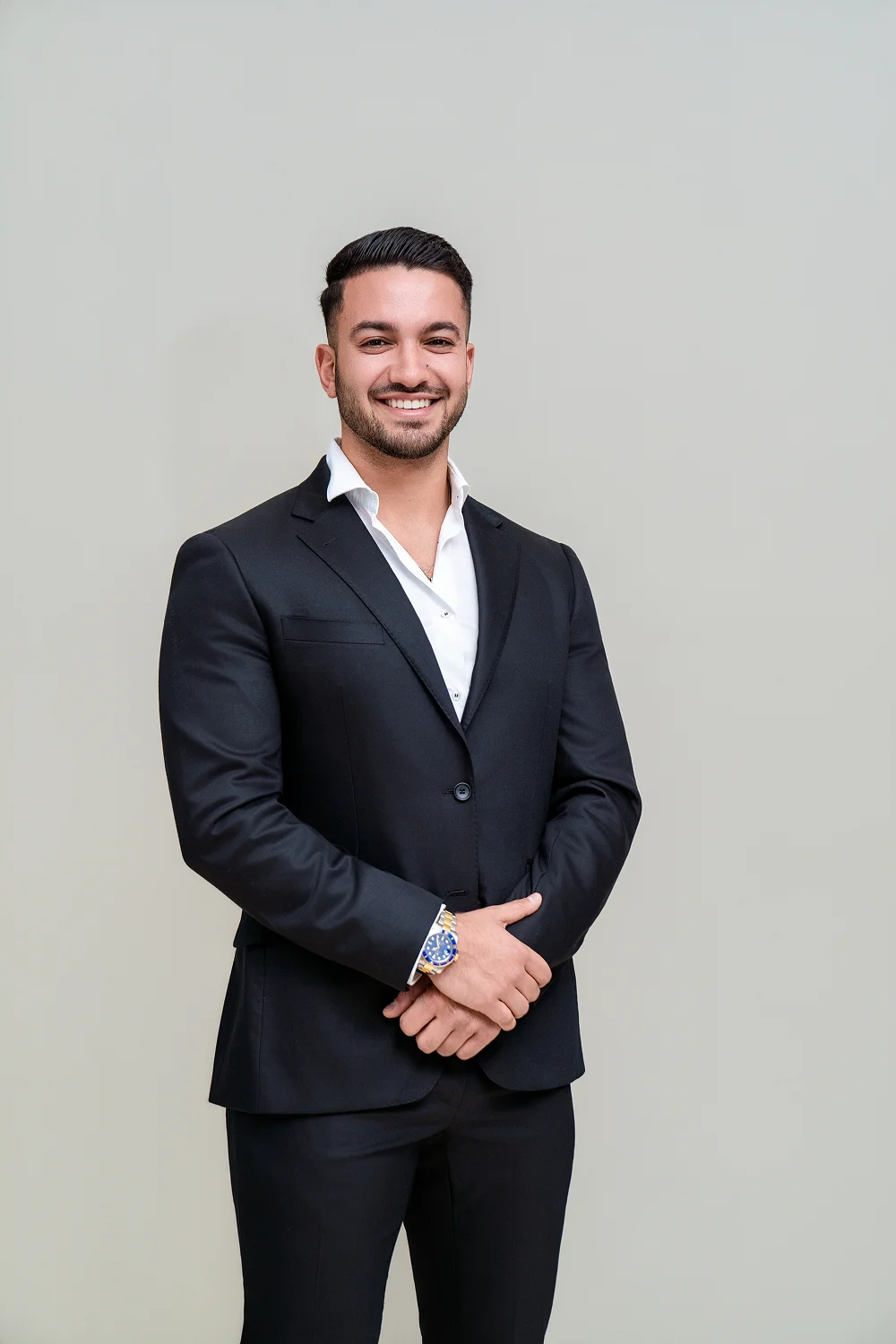 Smiling man in black suit and white shirt standing against a plain light gray background.