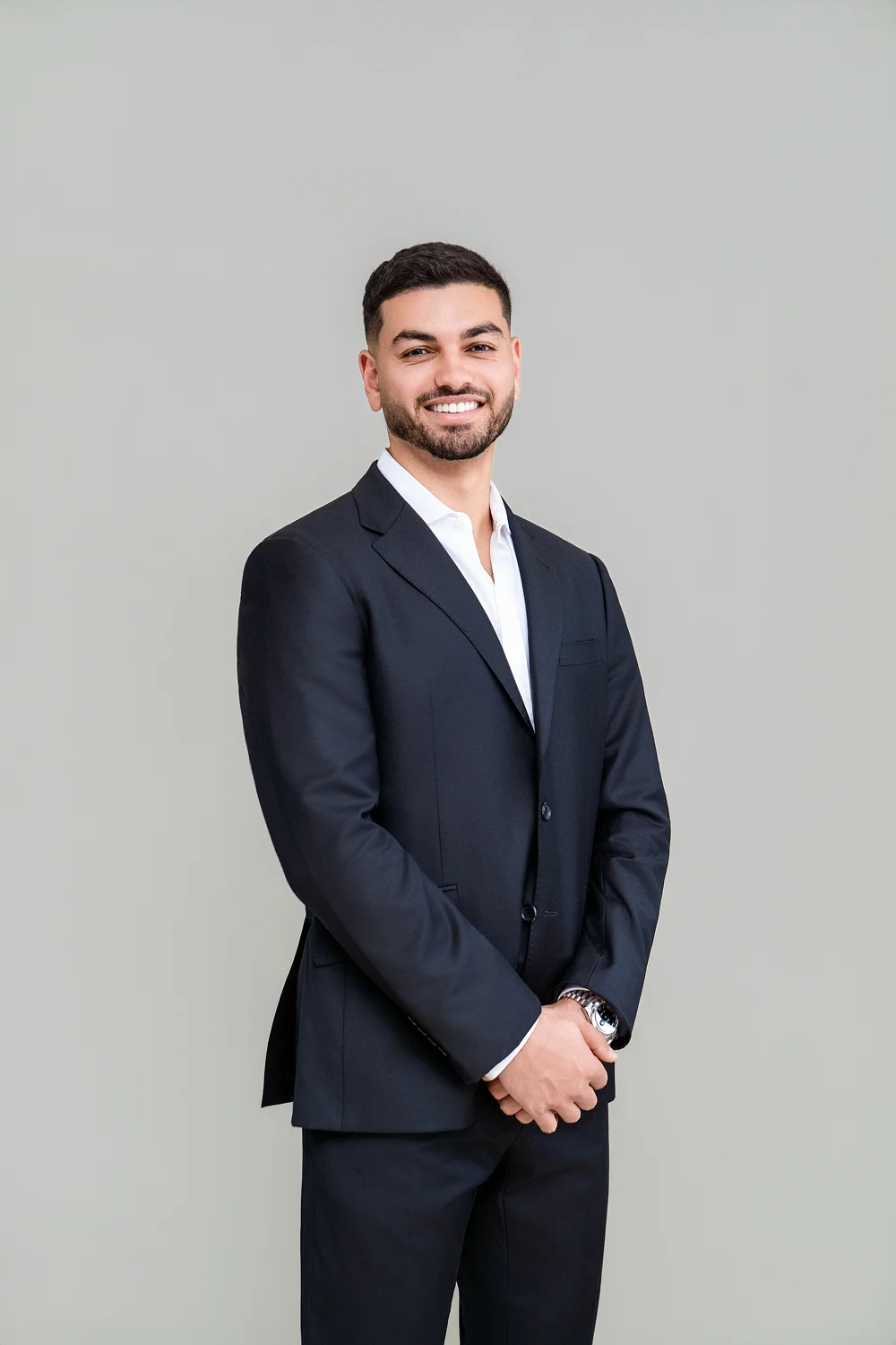 Smiling man in a dark suit and white shirt standing with hands clasped against a plain gray background.
