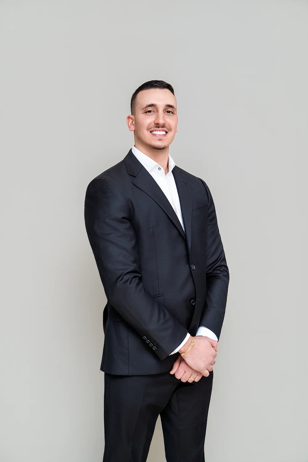 Smiling man in a black suit with a white shirt standing against a light gray background.