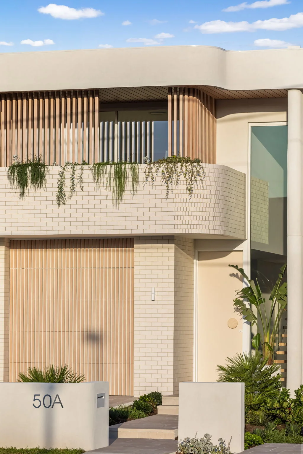 Modern two-story house with beige brick walls, wooden slats, hanging plants on balcony, and address 50A on white wall.