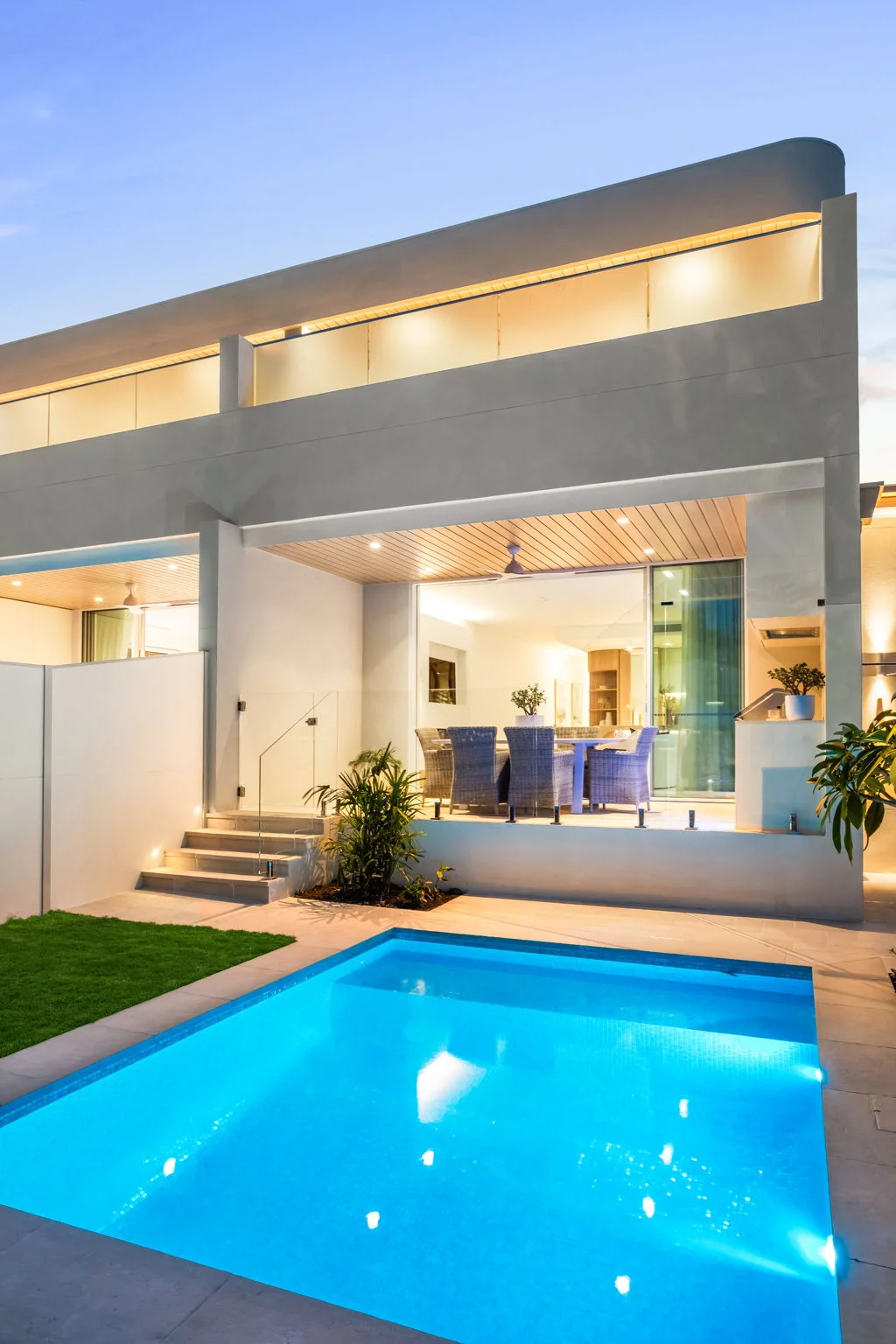 Modern house patio with dining table and chairs next to a lit blue swimming pool at dusk.