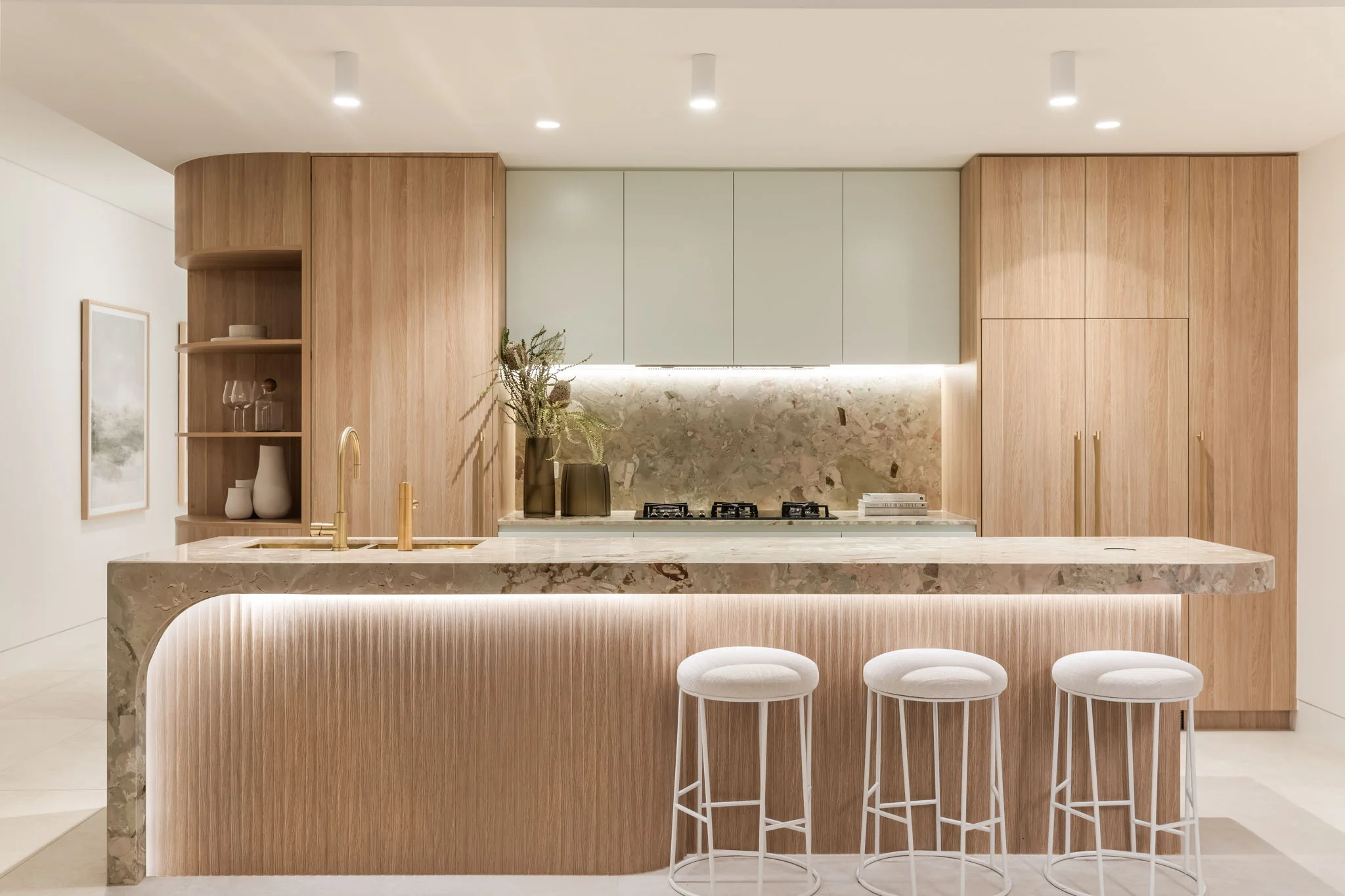 Modern kitchen with light wood cabinetry, marble countertop island with underlighting, gold faucet, and three white cushioned bar stools.