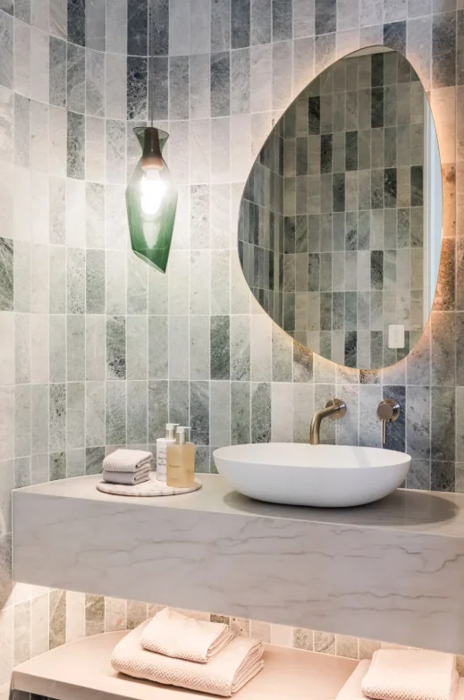 Modern bathroom vanity with white oval vessel sink, brass faucet, irregular oval backlit mirror, small towels, soap dispensers, and vertical grey green tiles covering walls.