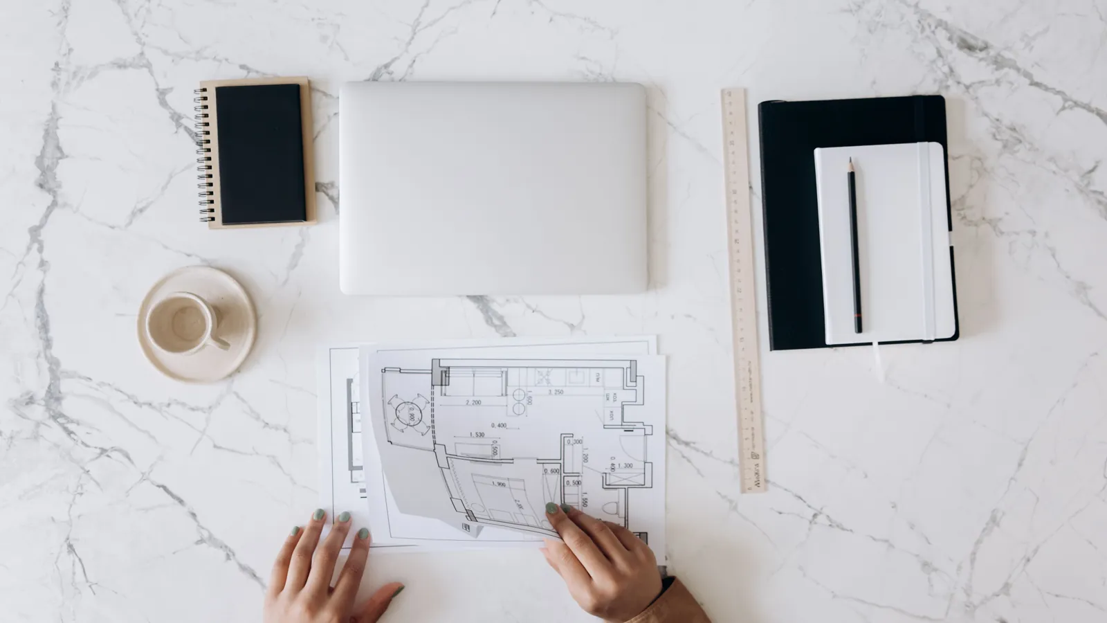 Overhead view of building plans, a laptop, notebooks and a ruler on a desk.