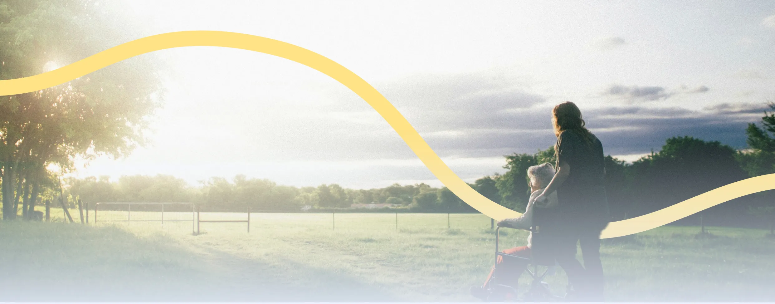 Person pushing an elderly woman in a wheelchair along a sunlit path near a green field with trees and cloudy sky.