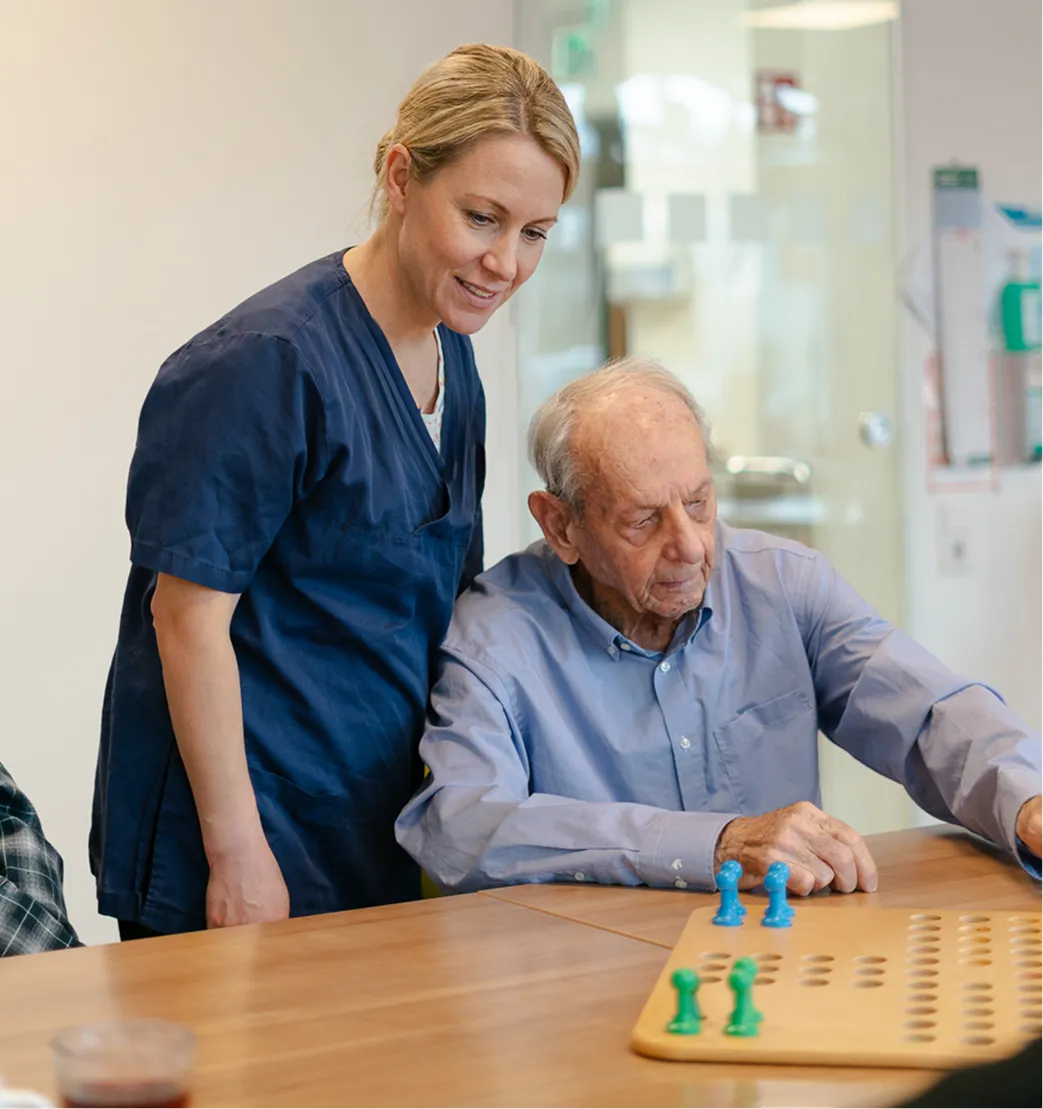 Elderly man playing a board game while a female caregiver in navy scrubs watches and supports him.