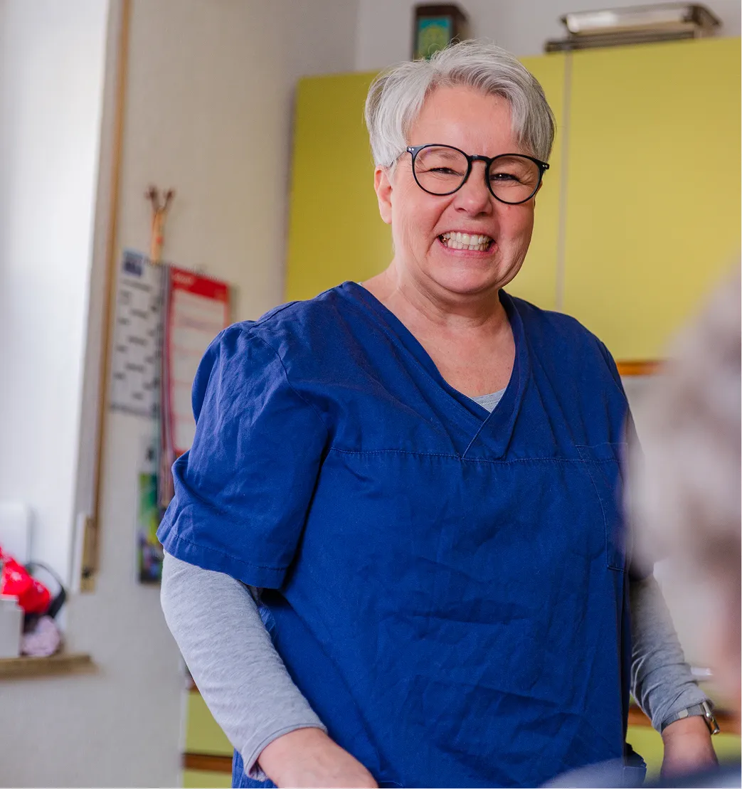Smiling older woman with short gray hair and glasses wearing blue medical scrubs and a gray long sleeve shirt underneath.