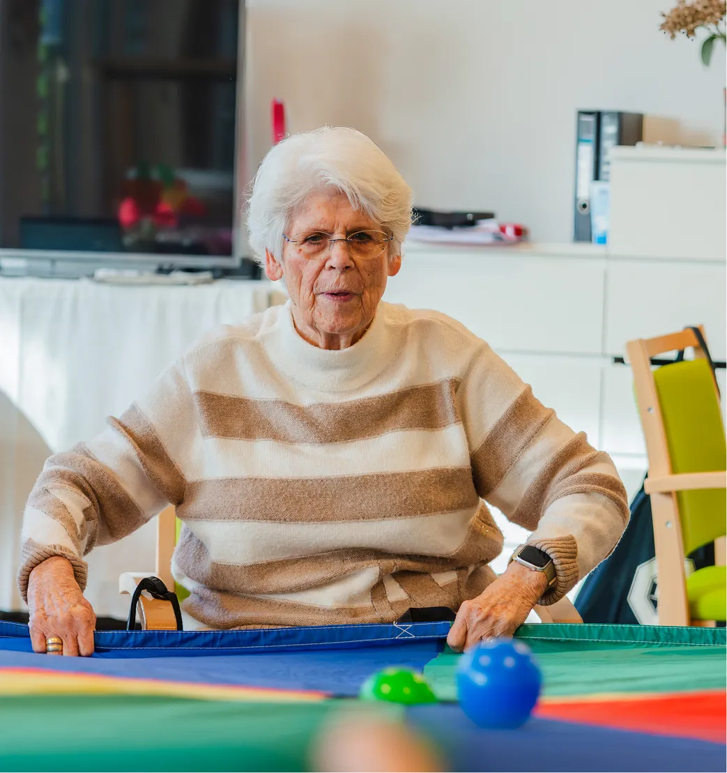 Elderly woman with white hair and glasses sitting at a colorful table holding a blue fabric edge indoors.