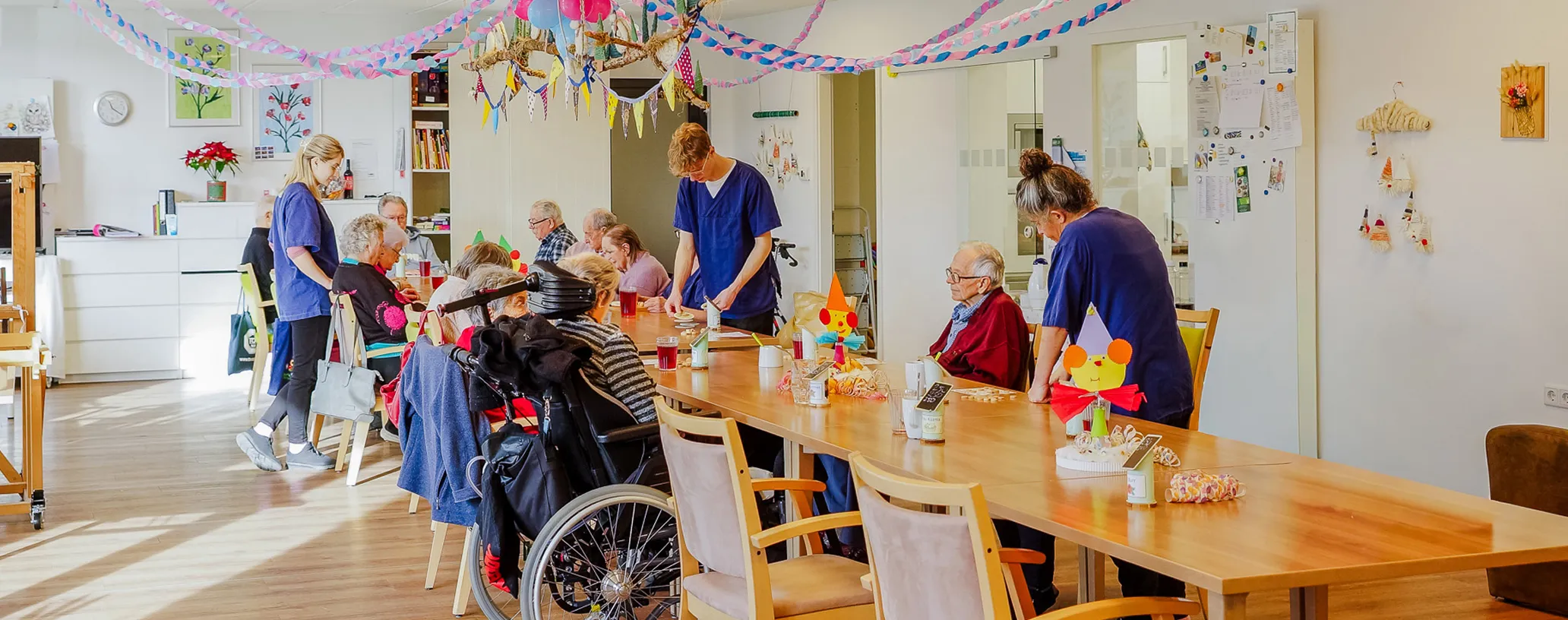 Elderly people seated around a decorated table in a bright room, being assisted by caregivers in blue uniforms.