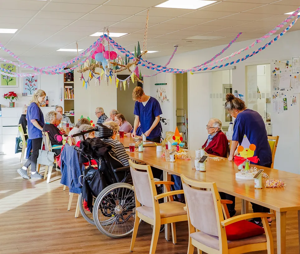 Elderly people and caregivers interacting around a long table decorated with colorful streamers and crafts in a bright communal room.