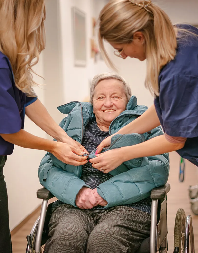Smiling elderly woman sitting in a wheelchair while two caregivers help button her teal winter coat.