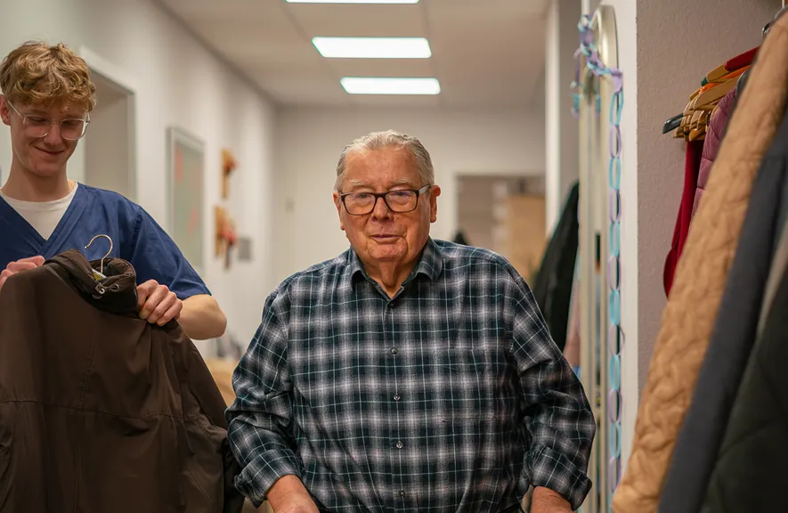 Elderly man in a plaid shirt standing in a hallway with a young man in scrubs holding a brown coat.