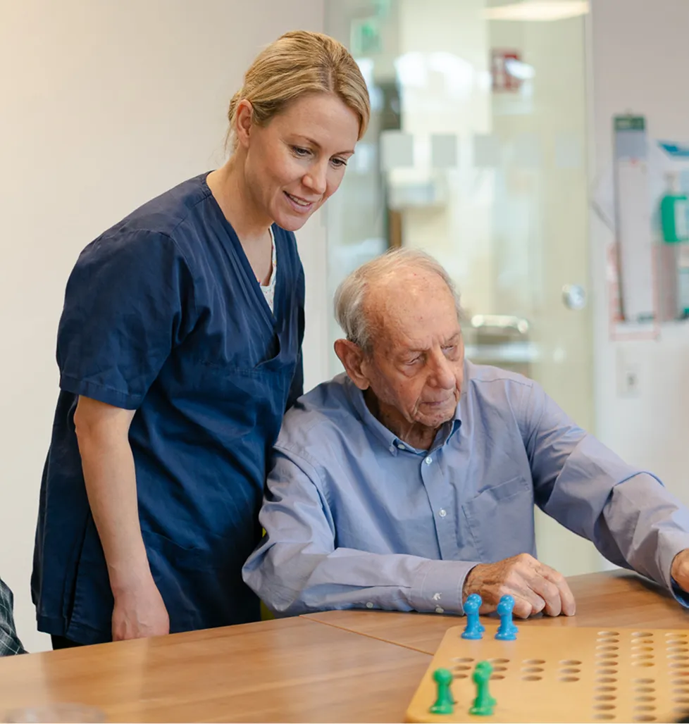 Elderly man playing board game with caregiver observing and smiling.
