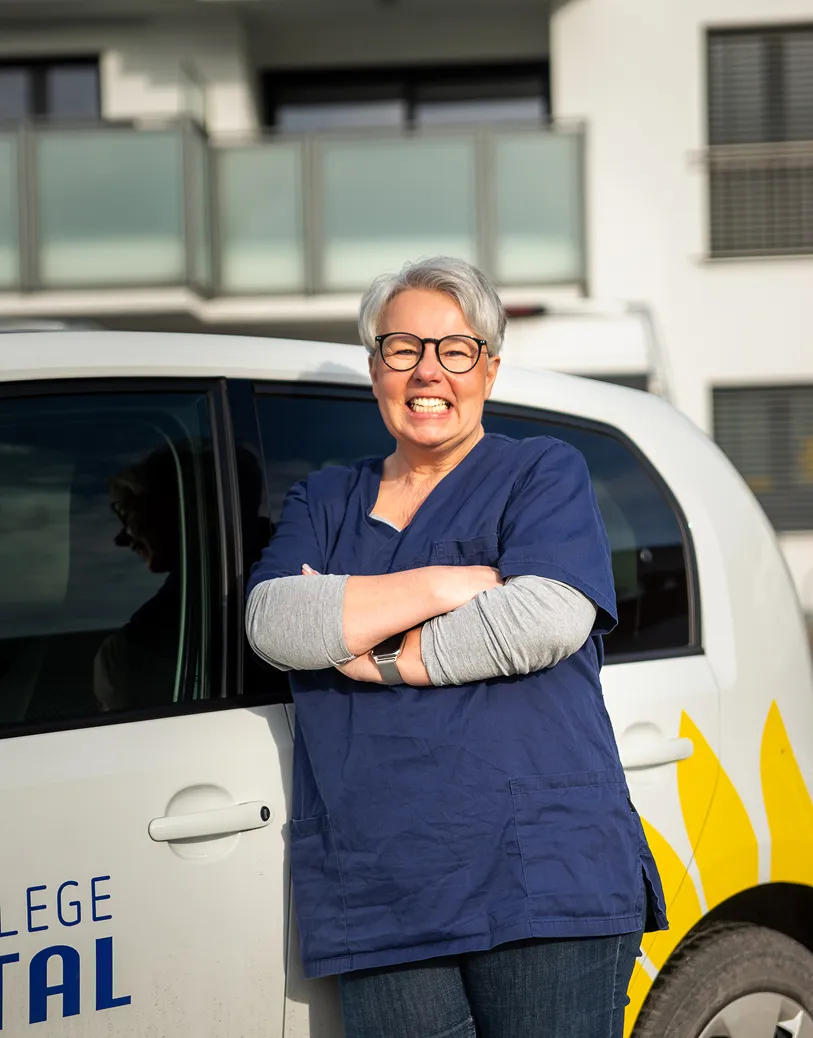 Smiling woman with short gray hair and glasses wearing navy scrubs standing with arms crossed in front of a white vehicle.