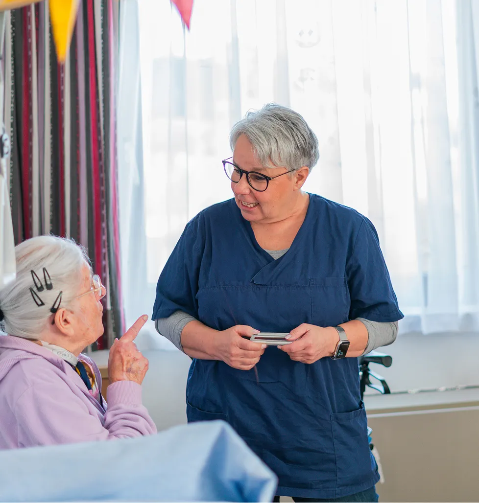 Healthcare worker in blue scrubs smiling and talking to an elderly woman in a lavender sweater in a well-lit room.