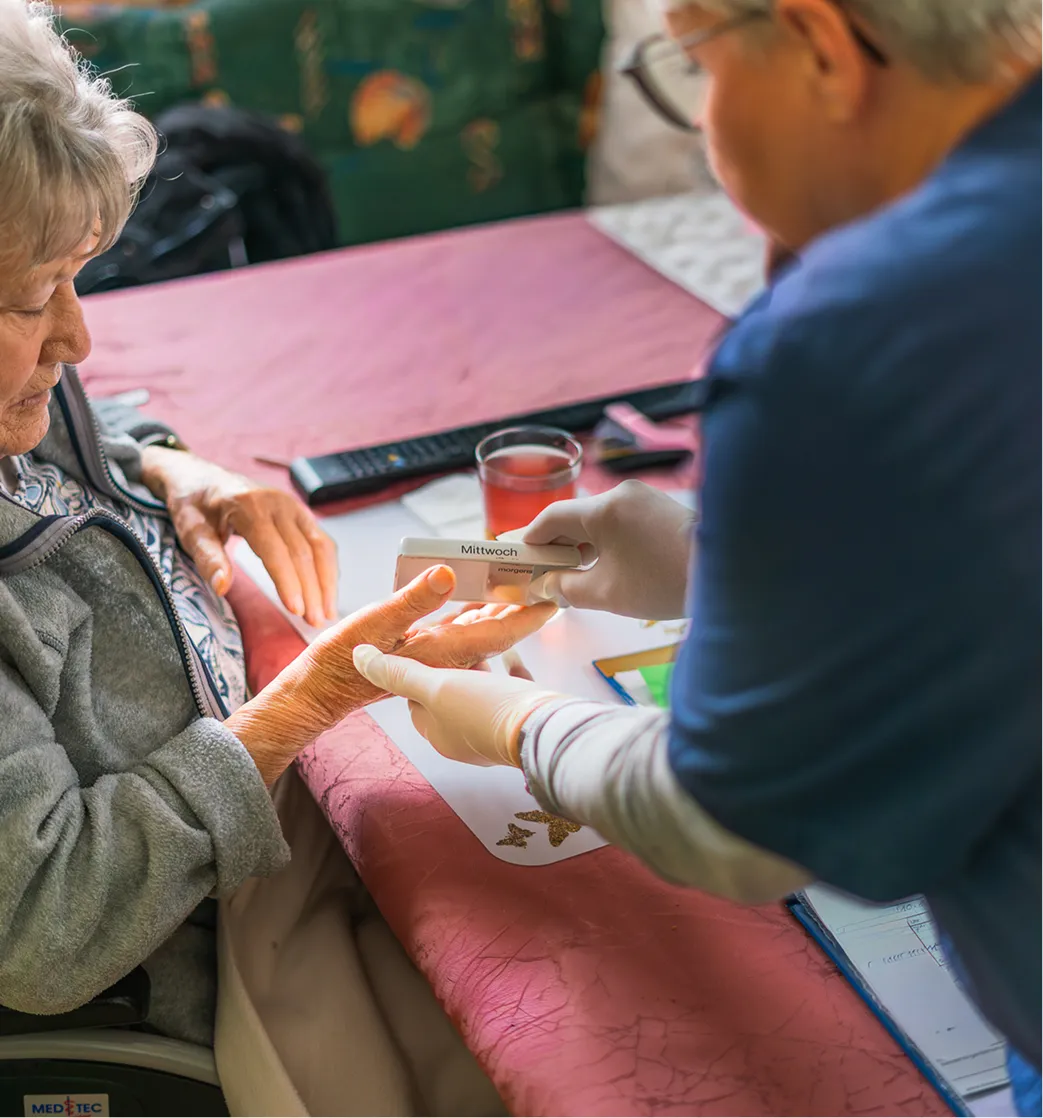 Caregiver wearing gloves assisting elderly woman by handing her a pill organizer labeled 'Mittwoch'.