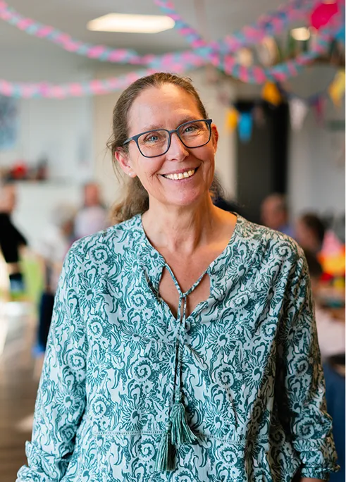 Smiling middle-aged woman with glasses wearing a patterned green blouse in a decorated indoor setting.