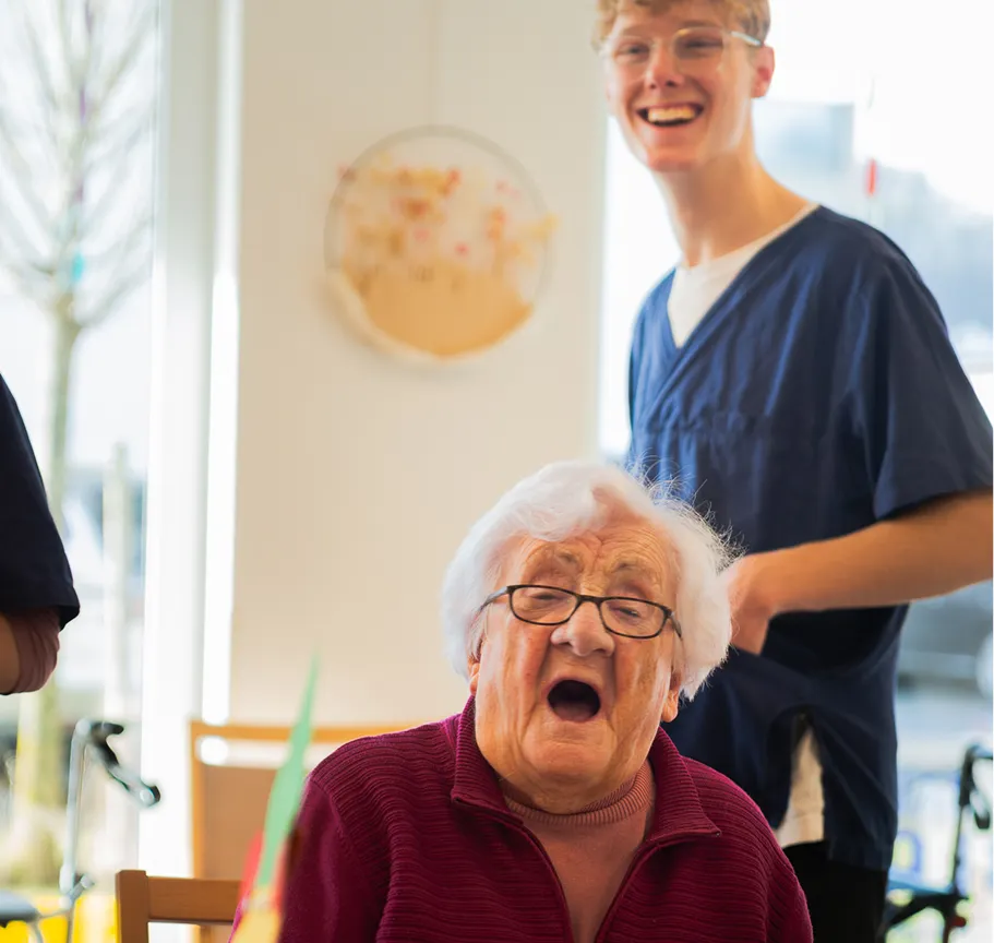 Elderly woman wearing glasses and a maroon jacket sitting and a young man in blue scrubs standing and smiling behind her in a bright room.