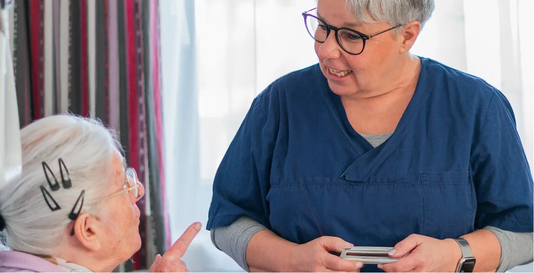Older woman with white hair and hair clips speaks while a healthcare worker in blue scrubs listens with a smile.