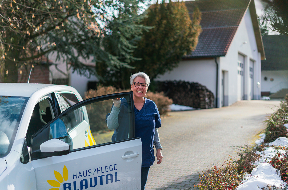 Smiling woman in blue uniform standing by an open car door with Hauspflege Blautal logo in a snowy residential area.