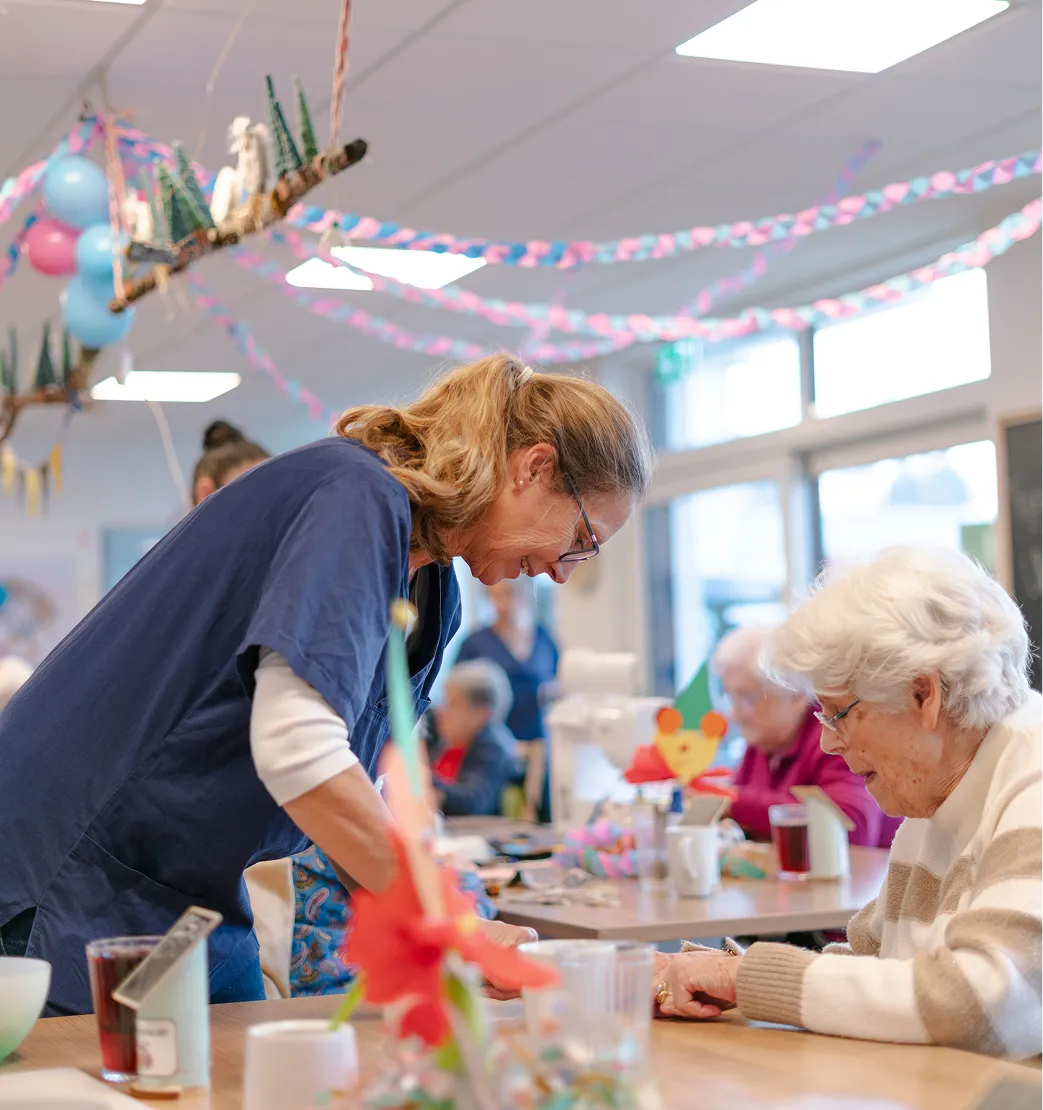 Caregiver in blue scrubs interacting warmly with an elderly woman at a decorated activity table in a senior care facility.