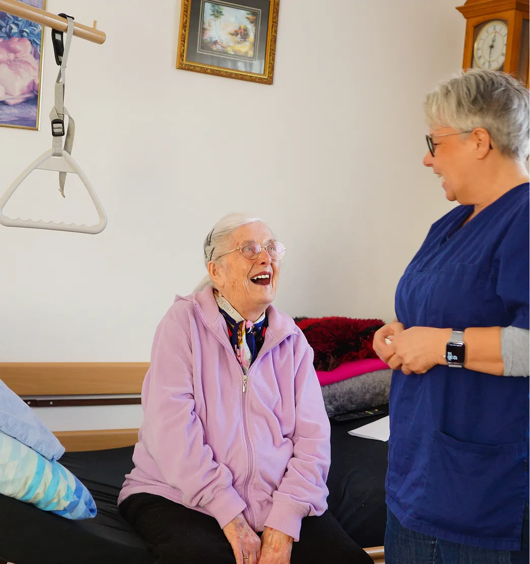 Elderly woman in a lavender jacket smiling and sitting on a bed talking with a female caregiver in navy scrubs.