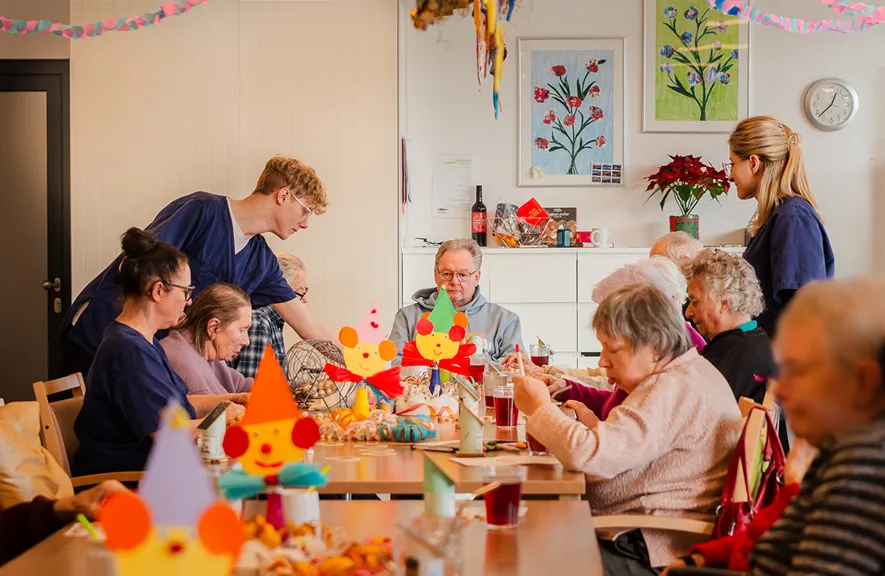 Elderly people sitting around a table with colorful party decorations, some engaging in activities, assisted by two caregivers in blue uniforms.