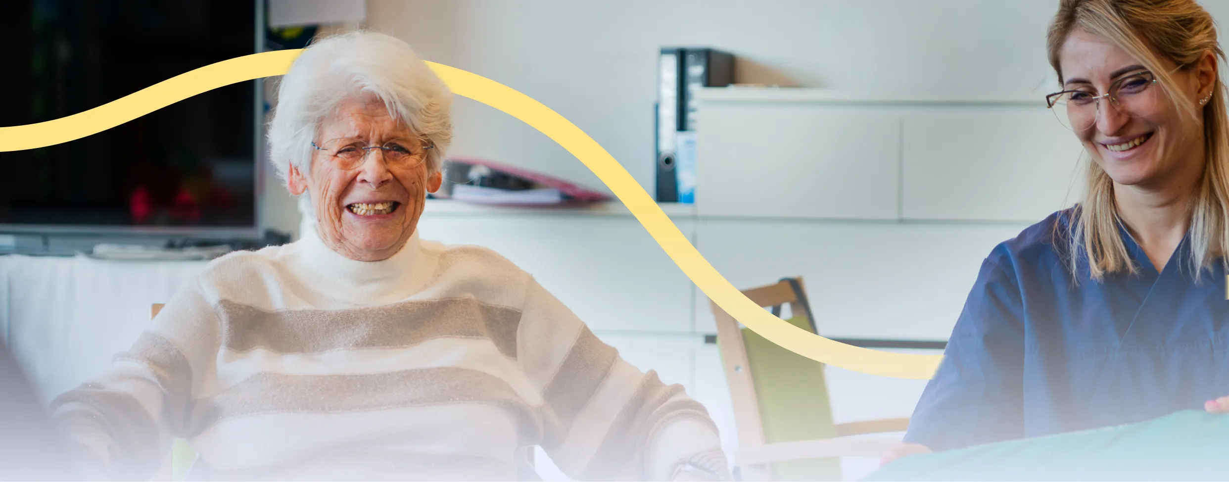 Caregiver in blue scrubs assisting and smiling with an elderly man indoors.