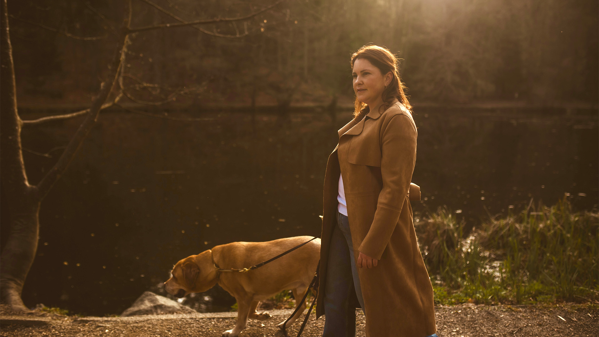 Terry Falk in braunem Mantel, die einen Hund an der Leine an einem ruhigen See mit herbstlichen Bäumen spazieren führt.